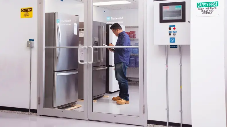 Our Senior Manager of Lab Operations, Jonathan Chan is standing in our refrigerator testing lab with a clipboard, jotting down test results. He's standing before a row of refrigerators. We're seeing him through the glass doors of the lab, which has a climate control box to its right.