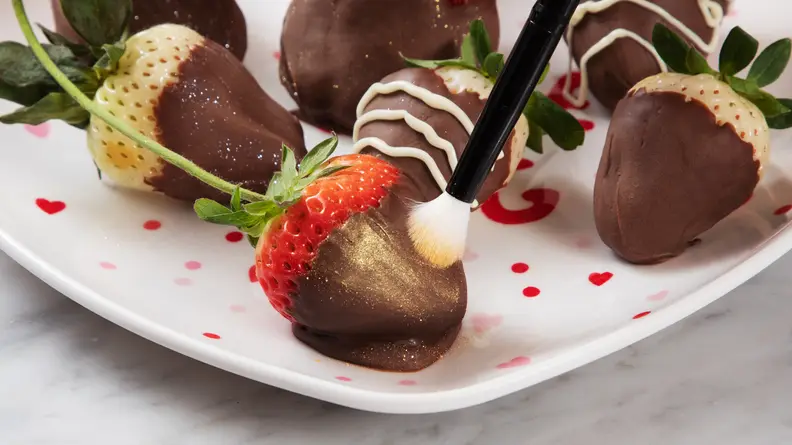 Closeup of a brush spreading gold edible glitter on a chocolate-covered strawberry.