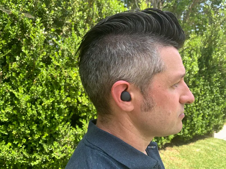 A man in a blue shirt with the Sony LinkBuds S in his ear stands in profile in front of a bush.