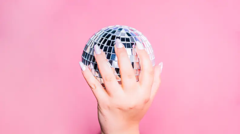 A hand wearing press-on nails holding a disco ball in front of a pink background.