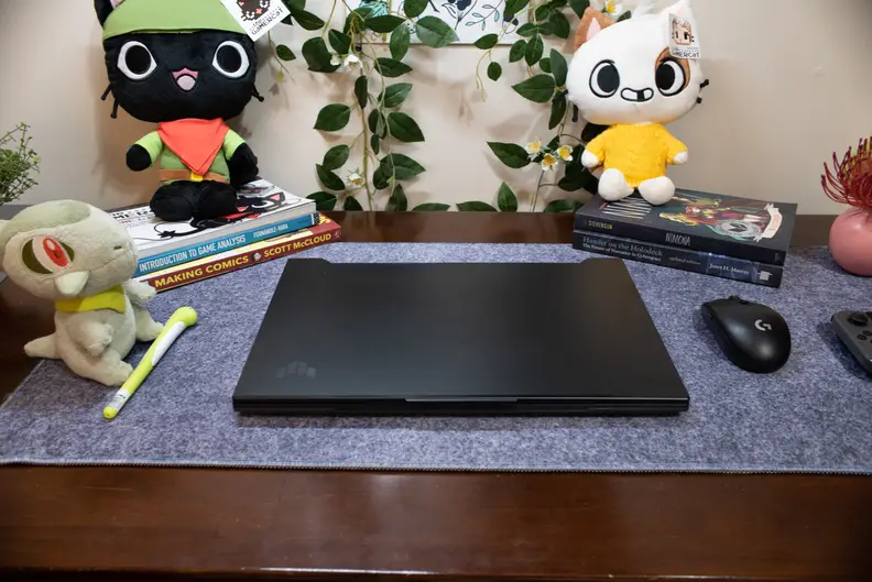 A closed, black laptop on top of a blue-gray mat with colorful plushies in the background