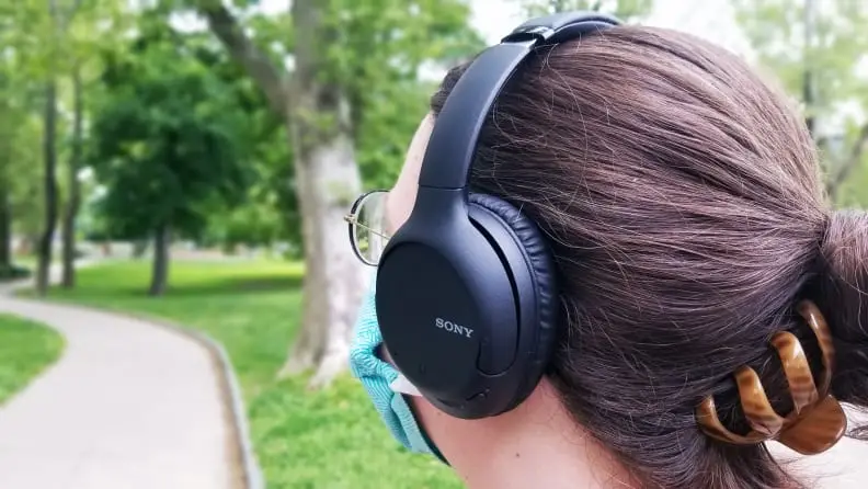 A person wearing a pair of Sony's WH-CH710N headphones with their hair in a bun and clipped, standing in a park near a walking path.