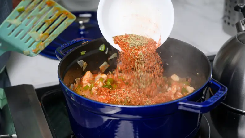 A bowl of spices and herbs being poured into a big pot of jambalaya cooking on a stove.