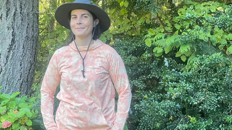 A woman wearing the Women's Solarfoil Pullover Hoodie and Trailcool UPF Cooling Sun Hat outside next to trees.