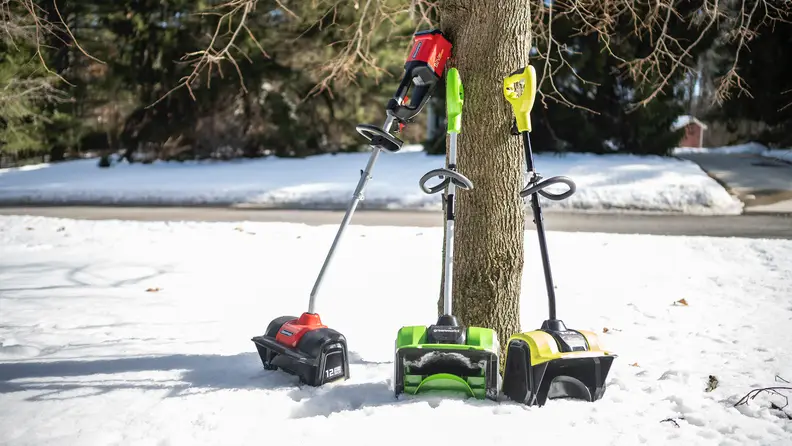 Three electric snow shovels lean up against a tree.