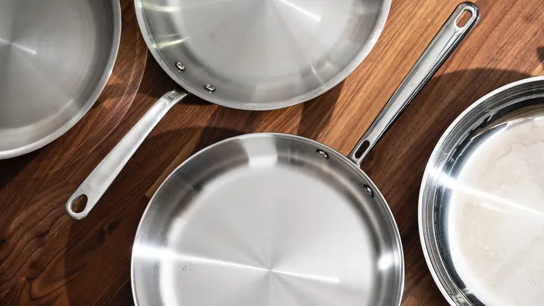 Four empty stainless steel skillets sit on a kitchen counter.