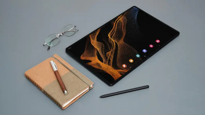 A Galaxy Tab 8 Ultra on a grey blue background with a notebook and pen, glasses, and stylus.