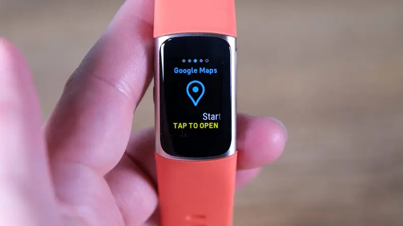 Person holding a Fitbit Charge 6 with an orange band has the Google Maps app on screen.