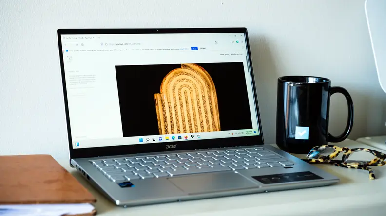 An open and powered on silver laptop with a black mug and a pair of glasses to its left and a brown notebook to its right.