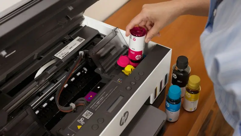 A close-up view of a person refilling the visible magenta tank on an HP Cartridge-free printer with a bottle.
