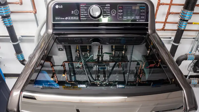 A close-up of an LG top-loader in our testing labs. Behind the appliance and in the reflection on its translucent lid you can see a network of pipes we use to regulate water temperature and flow.