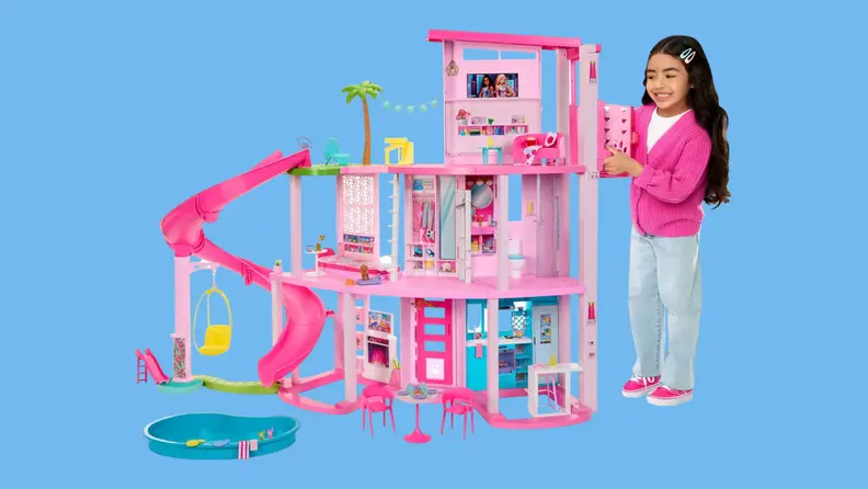 A young girl smiles as she stands next to the Barbie Dreamhouse 2023, with its three-story slide and pet play area.