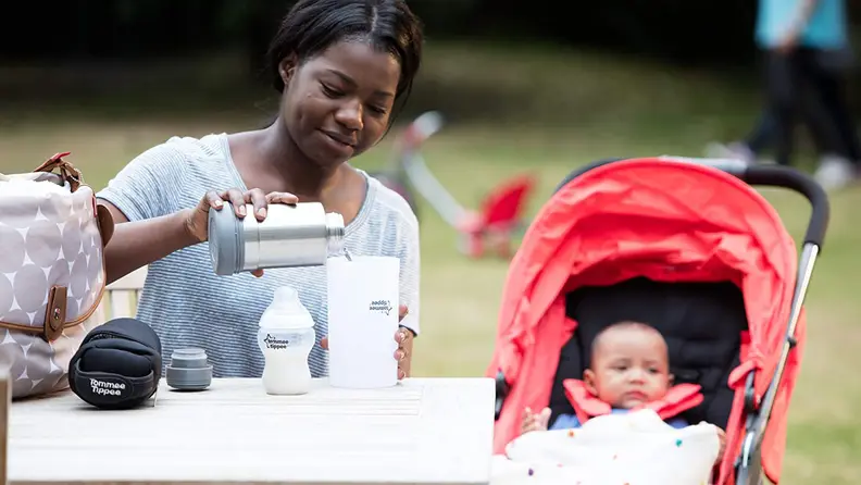 A mother using a portable bottle warmer on a picnic table with her baby next to her in a stroller.