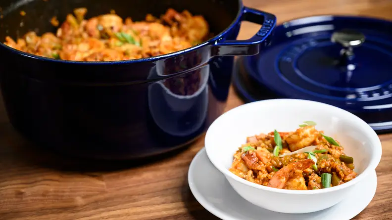 A pot full of jambalaya with the lid removed, next to a bowl full of the dish on a countertop.