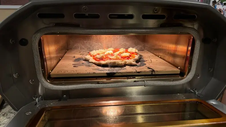 Raw pizza cooking inside of the Ooni Volt 12 pizza oven with door open.