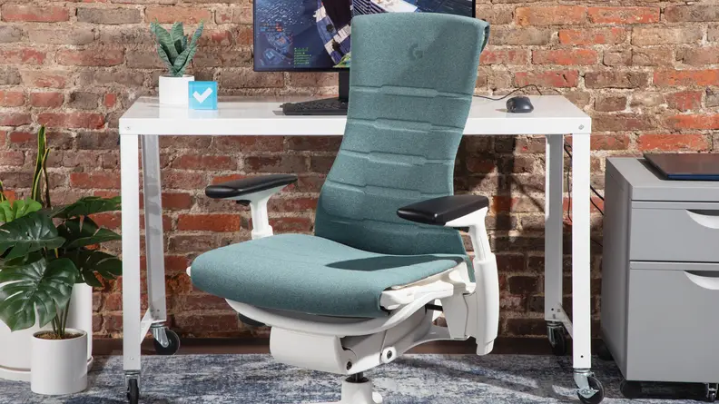 The Herman Miller Embody gaming chair in the color blue, next to a white office desk and office supplies.