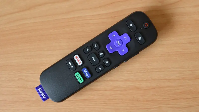 Roku Express 4K+ remote is all black with colorful buttons on top, including quick keys for streaming services like Netflix, Disney+, Hulu, and Apple TV+. It sits atop a yellow colored woodgrain table.