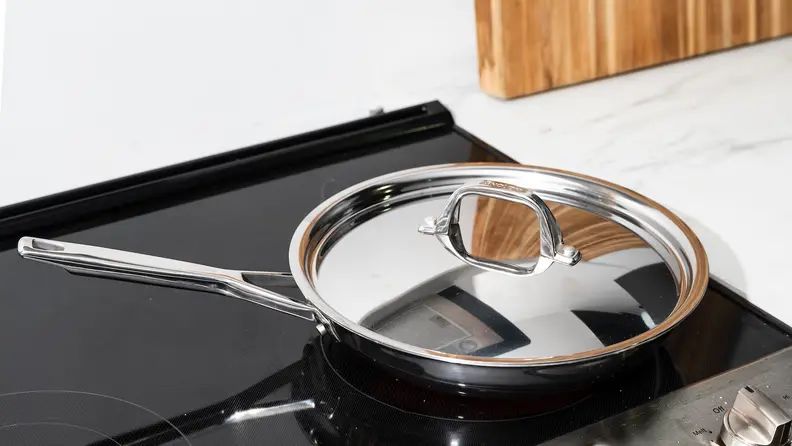 The Anolon Tri-Ply Clad Frying Pan with lid sits on a stove.