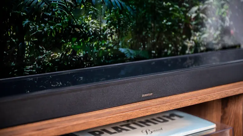 The glass-topped, all-black Bose 900 soundbar sits atop a wooden TV console with media beneath it and a TV behind it