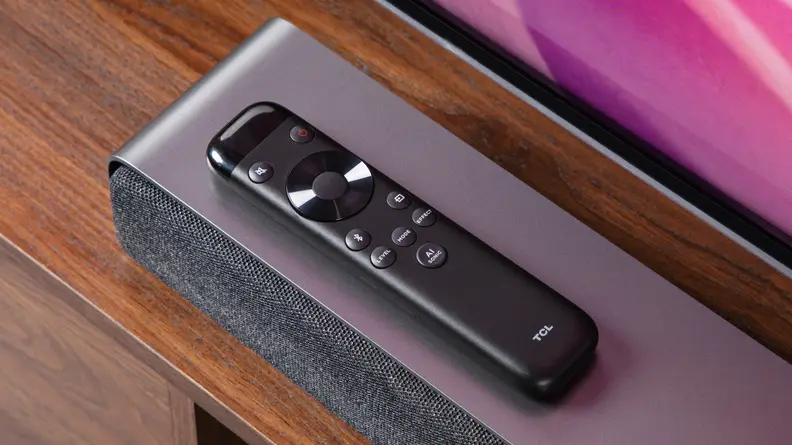 Small black remote sits on top of the TCL Q Series (Q6310) 3.1-Channel Soundbar that is on top of wooden surface.