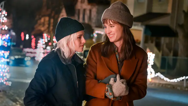 In Happiest Season (2020), Abby and Harper (Kristen Stewart and Mackenzie Davis) hold hands on a cold winter night as they walk down a street bundled in coats, hats, and gloves.