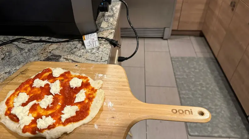 Raw pizza pie sitting on top of wooden pizza peel next to the Ooni Volt pizza oven.