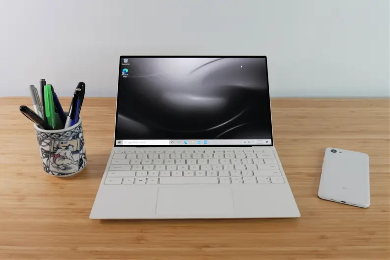 An open laptop on top of a desk with a cup of pens and a phone on either side.