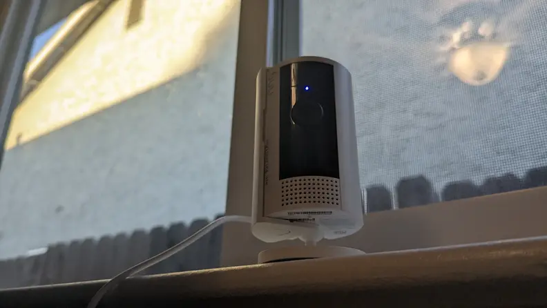 Shot from below of the 2nd-gen Ring Indoor Cam with camera light on while sitting on window sill in front of closed window on sunny day.