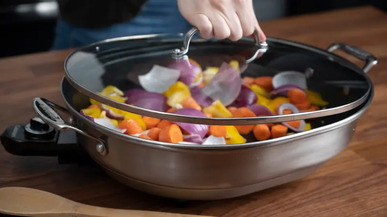Hand pulling the lid off of the Cuisinart Electric Skillet filled with vegetables.