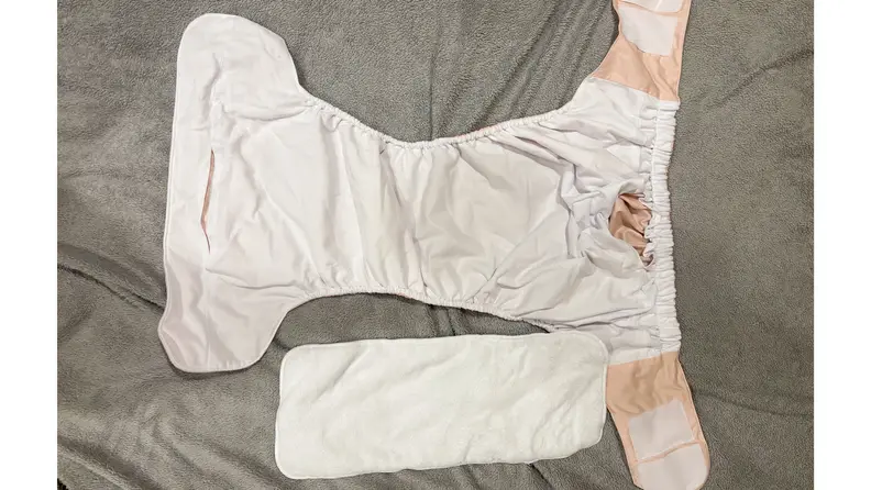A peach diaper with hook-and-loop closures laid open next to an insert on a gray fabric surface.