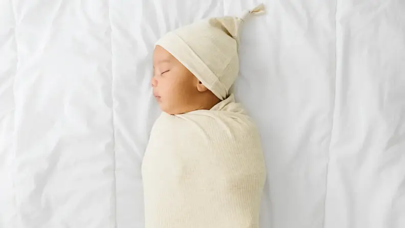 Swaddled baby peacefully sleeping on a white bed, wearing a cream-colored hat and wrap.