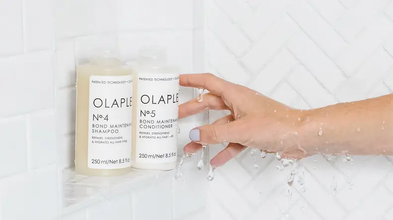A hand reaching toward two bottles of shampoo in a white shower stall.