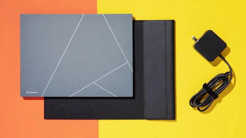 The Asus Zenbook S 13 OLED (2023) closed, laid out on an orange table, next to a black case and charger.