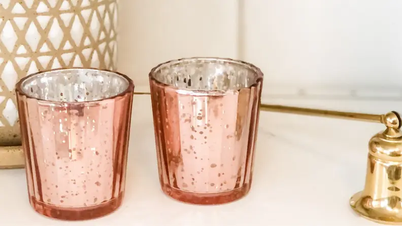 Two identical rose gold colored candles on white counter, next to gold bell and green potted plant.
