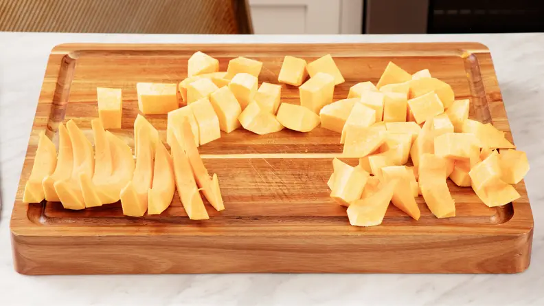 Butternut squash sitting on top of wooden cutting board after being cut into various shapes before being cooked.
