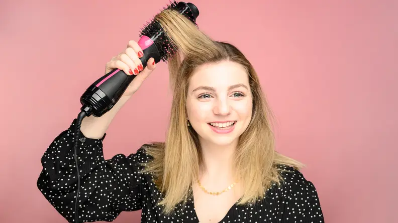 A woman running a hair dryer brush through her blond hair.