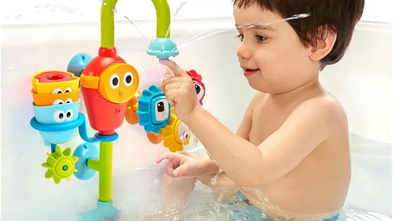 Toddler child plays with a vibrantly colored waterpark-style bath toy that suctions to the wall and sprays water.