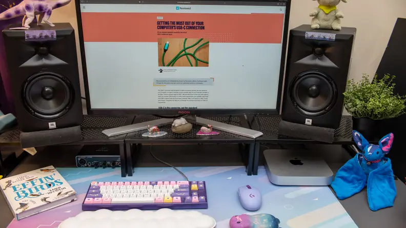 Desktop set-up featuring the Apple Mac Mini M2, a desktop computer, two speakers, a colorful computer keyboard, a plush stuffed animal and a book.