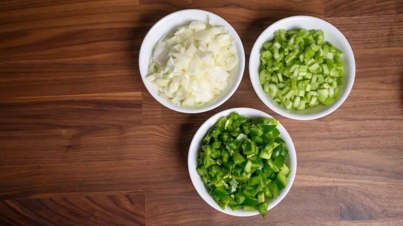 Three small white bowls of bell pepper, onion, and celery arranged together on a countertop, shot from above.