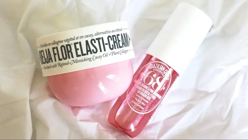 A pink bottle and tub of body lotion and spray on a sheet.