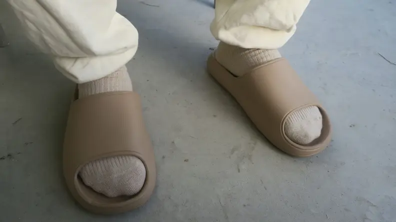 A pair of feet standing in beige pool slide sandals from Litfun, in off-white socks and cream colored sweatpants.