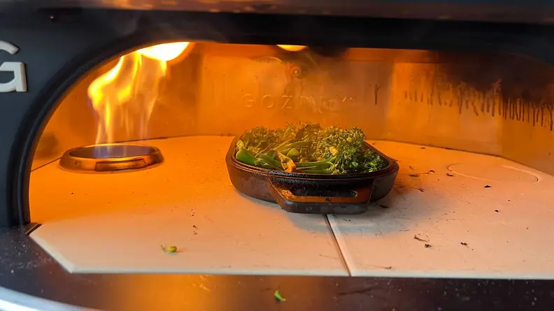 Broccolini cooking in cast-iron dish inside of Gozney Dome Pizza Oven with open flame.