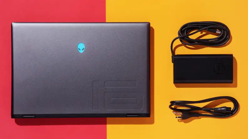 A closed Alienware m16 R2 gaming laptop next to a charging box and power cord on top of a red and yellow surface.