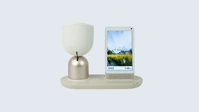 A white-hued ElliQ on its charging stand in front of a light blue background.