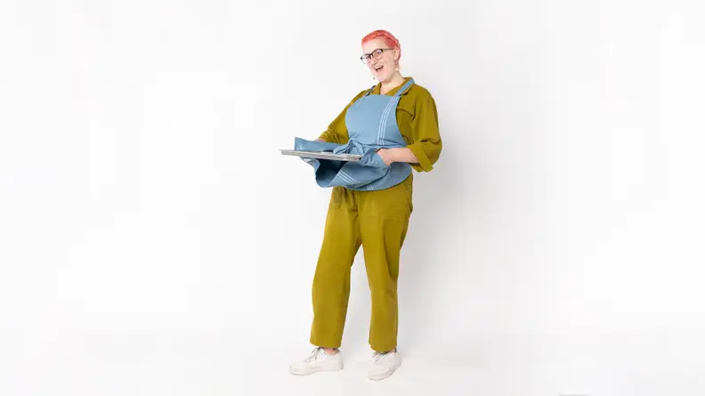 A person wearing a blue apron with built-in potholders, holding a metal sheet pan.