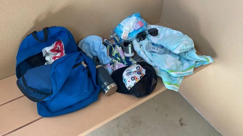 A backpack, towel, sunglasses, baby wipes, water bottle, and other items on a bench