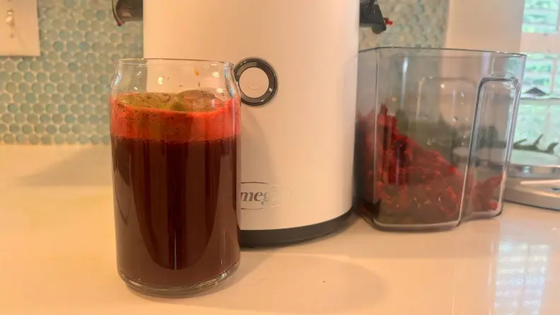 A glass of dark red pressed juice in front of the Omega Effortless Batch Juicer and a container of fruit.