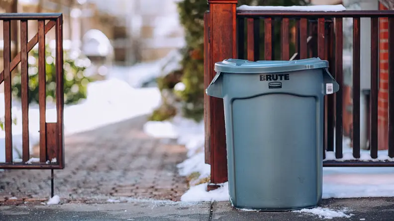 A grey Rubbermaid Brute outdoor trash can on a sidewalk in front of an open fence.