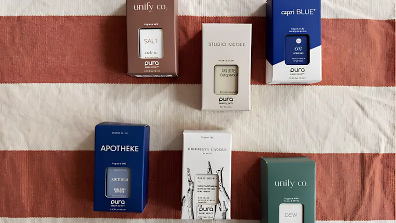 Pura diffuser review showing boxed fragrance refills from brands including Capri Blue, Apotheke, and Unify Co. arranged on a striped fabric background.