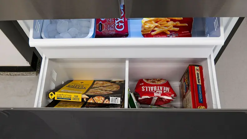 A close-up of the fridge's open freezer drawer, showcasing its main bin and the shelf that sits above it. The left corner of the upper shelf houses an ice reservoir. The rest of the space is filled with frozen foods.